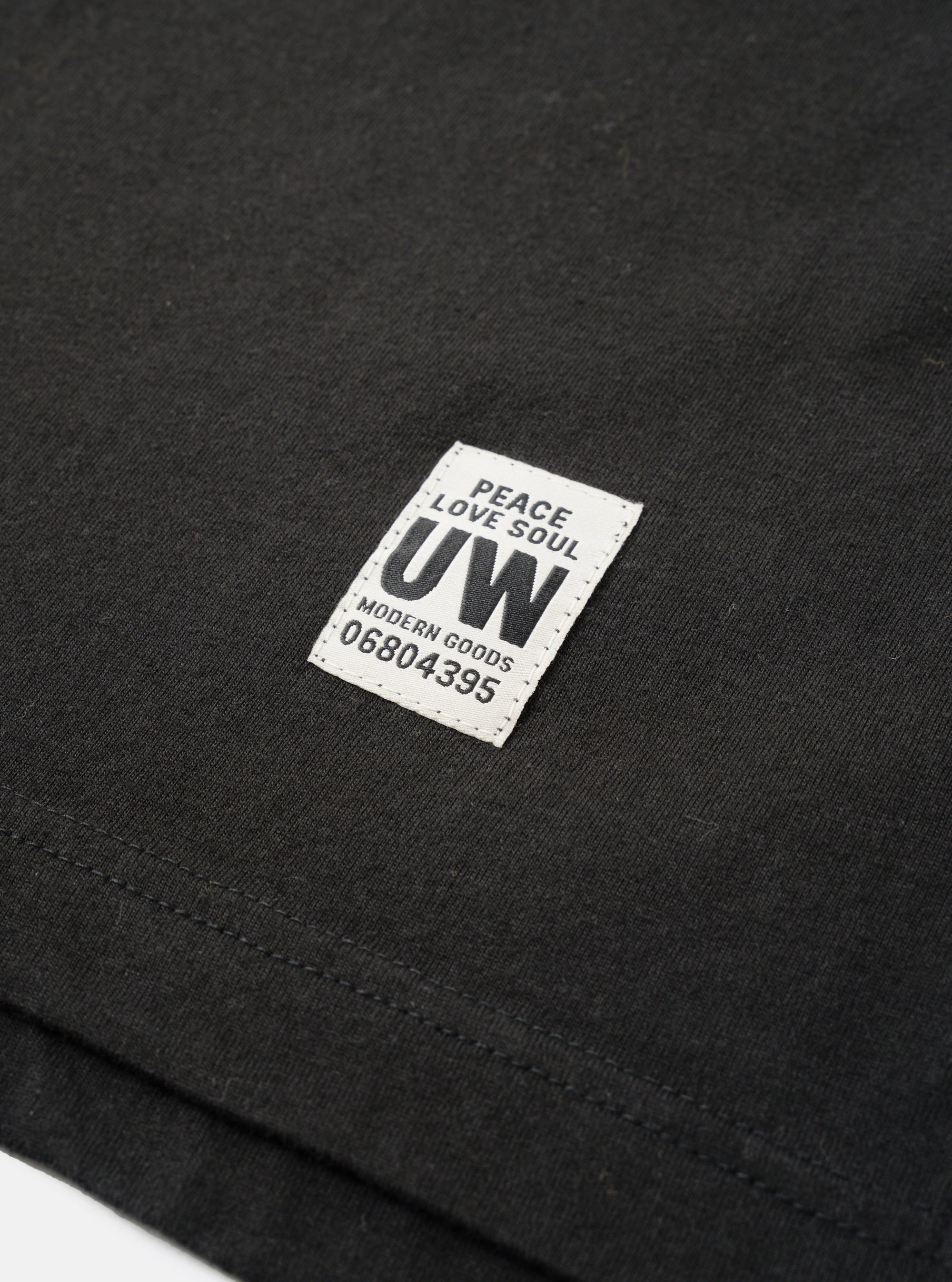 Close-up detail shot of a black long-sleeve pocket tee in single jersey cotton. A rectangular woven label reading "Peace Love Soul UW Modern Goods 06804395" is stitched onto the chest pocket. The hem and pocket stitching are visible. Shot as a flat lay on a white surface.