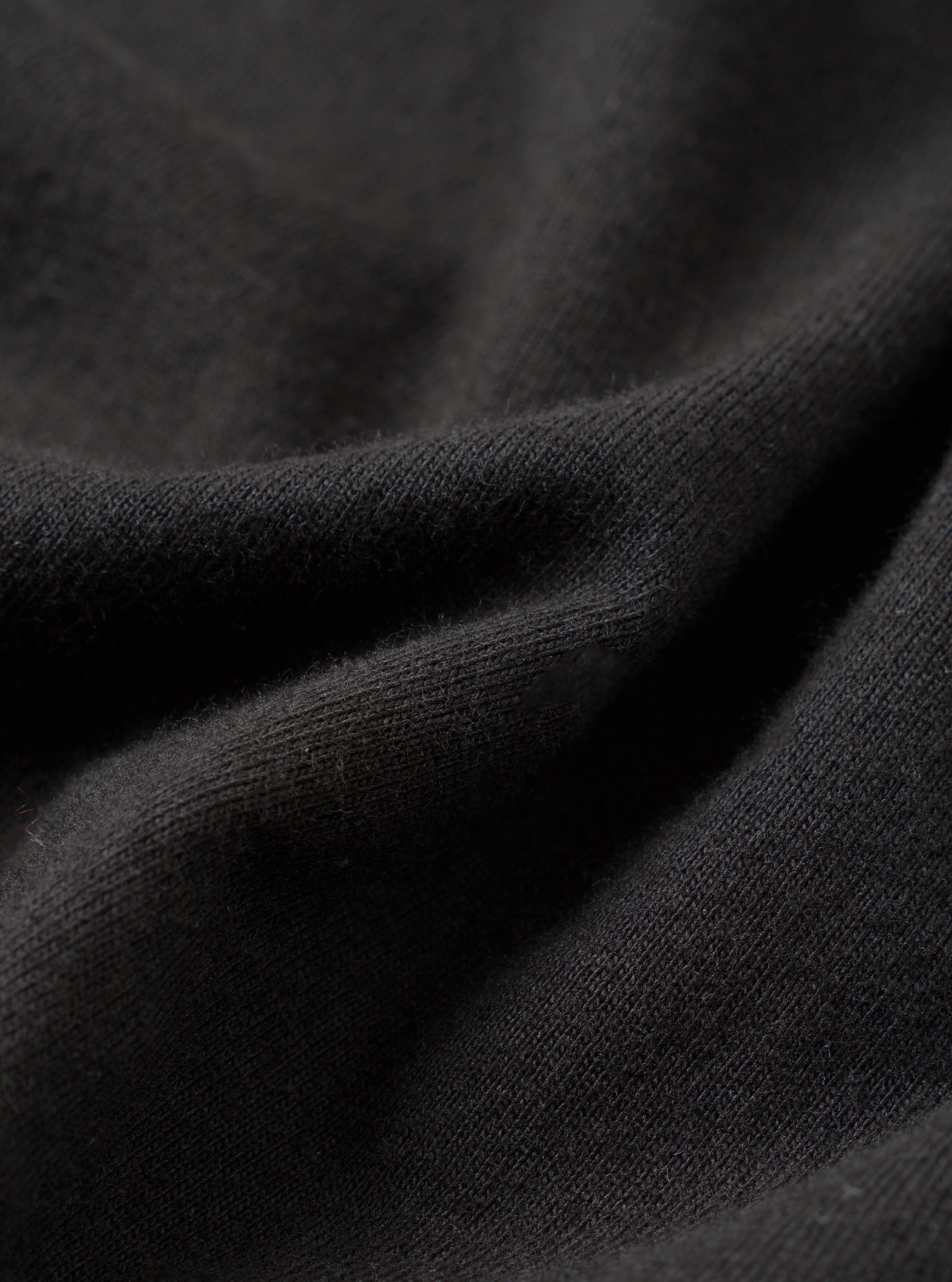 Close-up detail shot of a black single jersey knit fabric, showing soft draping folds with a fine, smooth knit texture visible at the ridges. The surface has a slightly brushed, matte finish. No garment details or background are distinguishable at this macro scale.