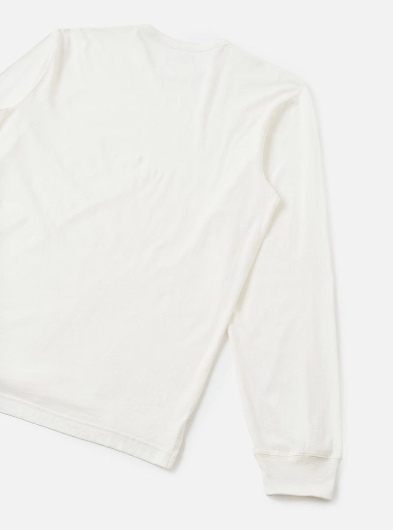 Flat lay of an ecru long-sleeve pocket tee on a light grey seamless backdrop. The shot is cropped to show the back and one full sleeve. The fabric is smooth single jersey cotton with natural drape. A ribbed cuff finishes the sleeve hem.