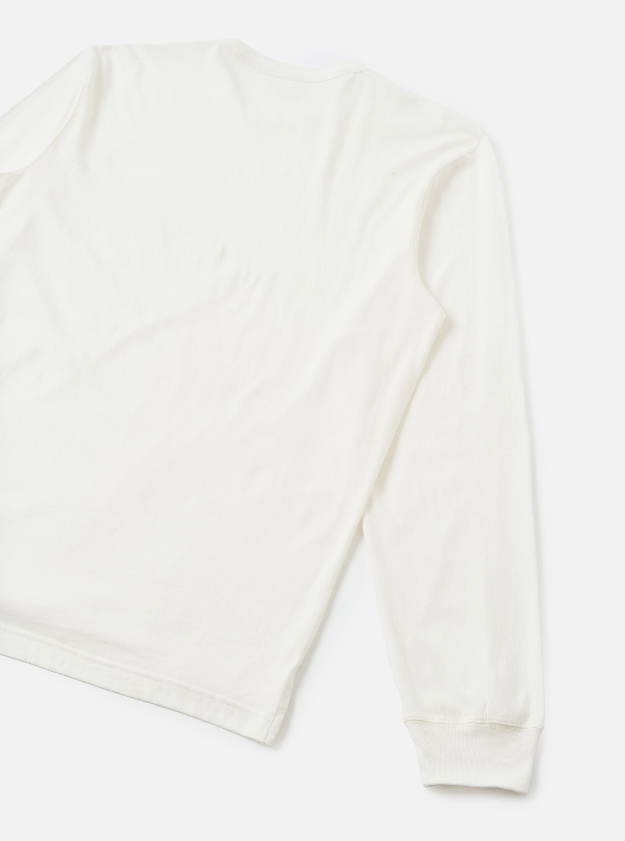 Flat lay of an ecru long-sleeve pocket tee on a light grey seamless backdrop. The shot is cropped to show the back and one full sleeve. The fabric is smooth single jersey cotton with natural drape. A ribbed cuff finishes the sleeve hem.