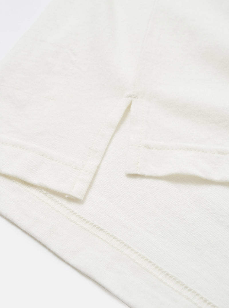Close-up detail of an ecru long-sleeve pocket tee in single jersey cotton. The shot shows the chest patch pocket with a clean folded top edge, alongside the hemmed cuff of the sleeve. Topstitching is visible along the hem. Flat lay on a white surface. No pattern, plain fabric with a fine, slightly textured jersey weave.