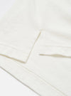 Close-up detail of an ecru long-sleeve pocket tee in single jersey cotton. The shot shows the chest patch pocket with a clean folded top edge, alongside the hemmed cuff of the sleeve. Topstitching is visible along the hem. Flat lay on a white surface. No pattern, plain fabric with a fine, slightly textured jersey weave.
