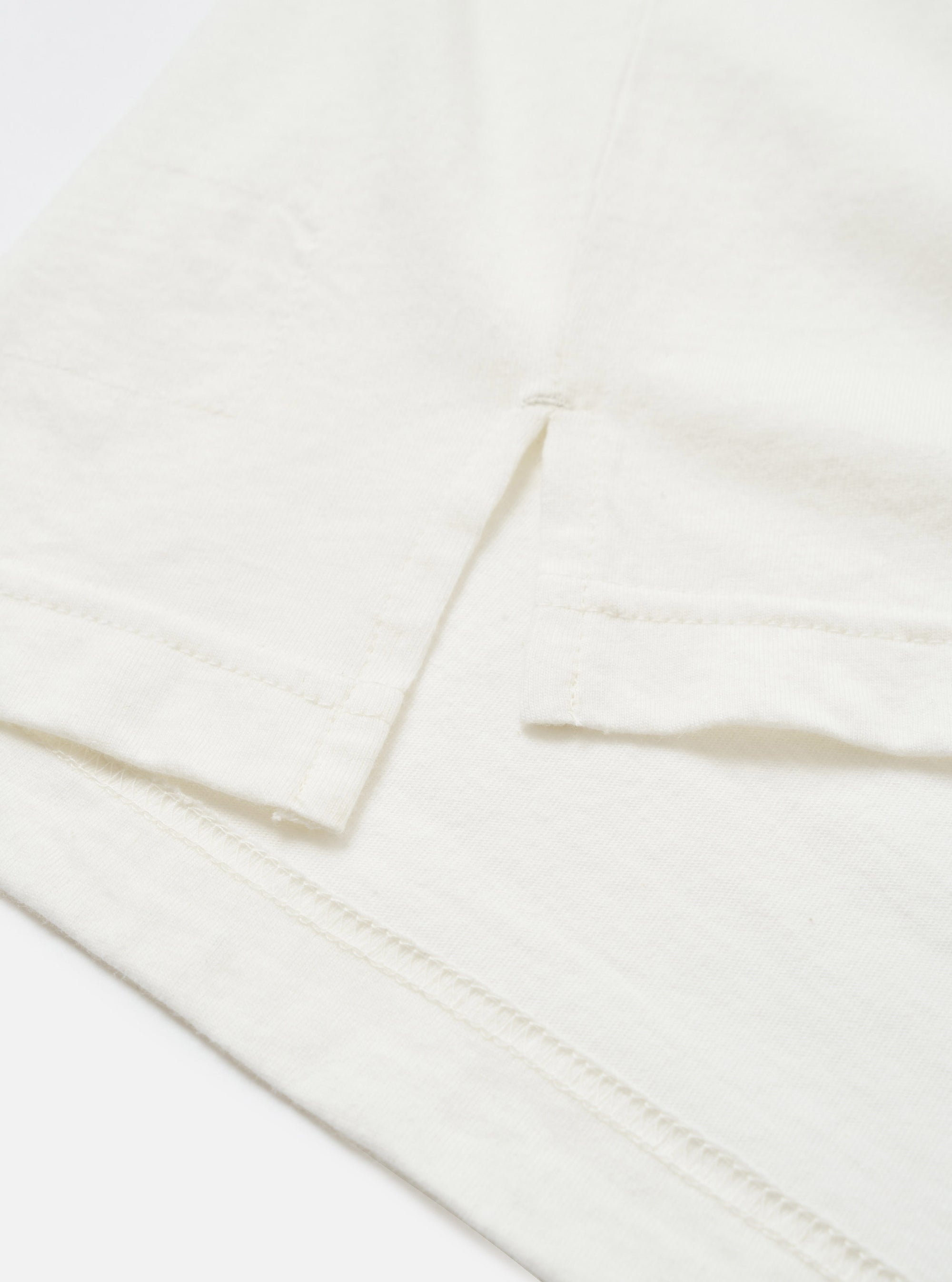 Close-up detail of an ecru long-sleeve pocket tee in single jersey cotton. The shot shows the chest patch pocket with a clean folded top edge, alongside the hemmed cuff of the sleeve. Topstitching is visible along the hem. Flat lay on a white surface. No pattern, plain fabric with a fine, slightly textured jersey weave.
