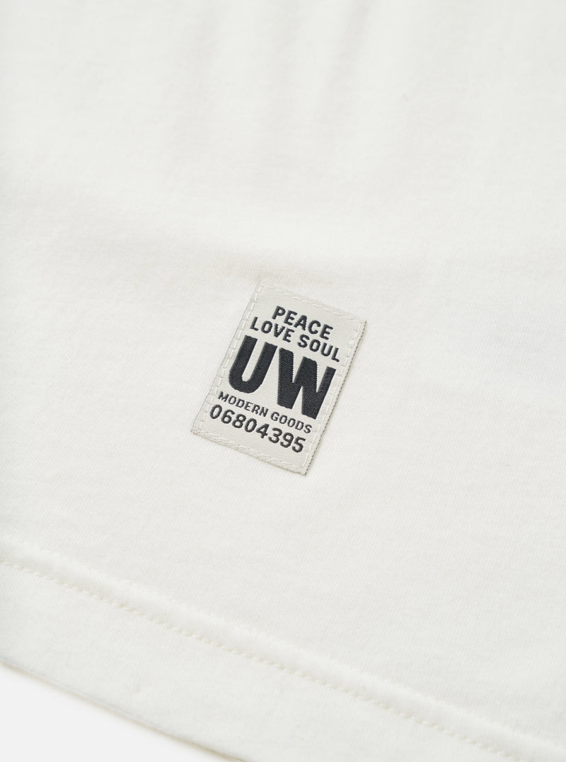 Close-up detail shot of an ecru single jersey long-sleeve pocket tee, showing a woven rectangular badge reading "Peace Love Soul, UW, Modern Goods, 06804395" in navy text on a light grey background. The hem seam is visible at the bottom of the frame. Plain white studio backdrop.