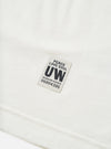 Close-up detail shot of an ecru single jersey long-sleeve pocket tee, showing a woven rectangular badge reading "Peace Love Soul, UW, Modern Goods, 06804395" in navy text on a light grey background. The hem seam is visible at the bottom of the frame. Plain white studio backdrop.