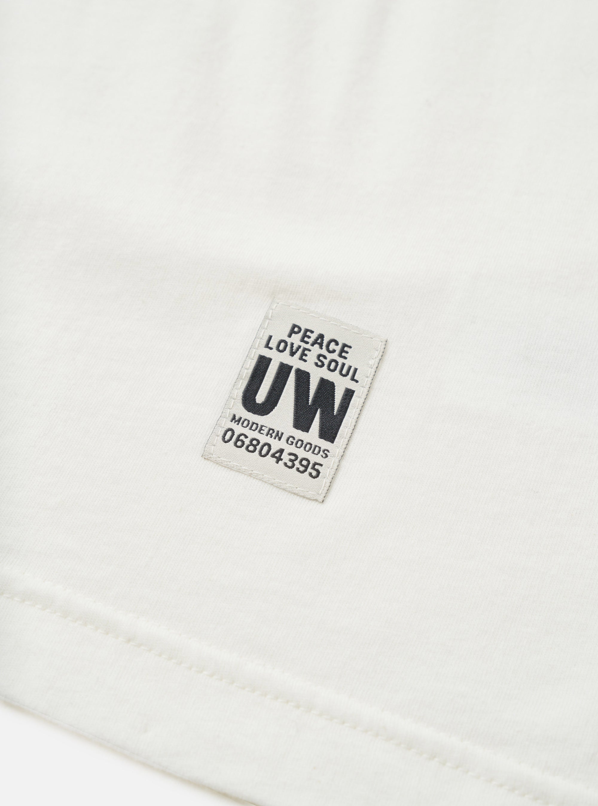 Close-up detail shot of an ecru single jersey long-sleeve pocket tee, showing a woven rectangular badge reading "Peace Love Soul, UW, Modern Goods, 06804395" in navy text on a light grey background. The hem seam is visible at the bottom of the frame. Plain white studio backdrop.