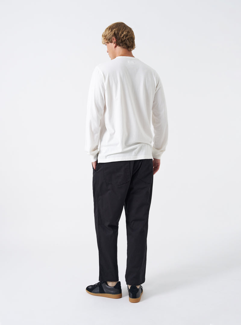 Back view of a model wearing the Ecru Single Jersey L/S Core Pocket Tee against a plain light background. The shot displays the off-white long-sleeve tee from behind, emphasizing the fit across shoulders and clean hemline.
