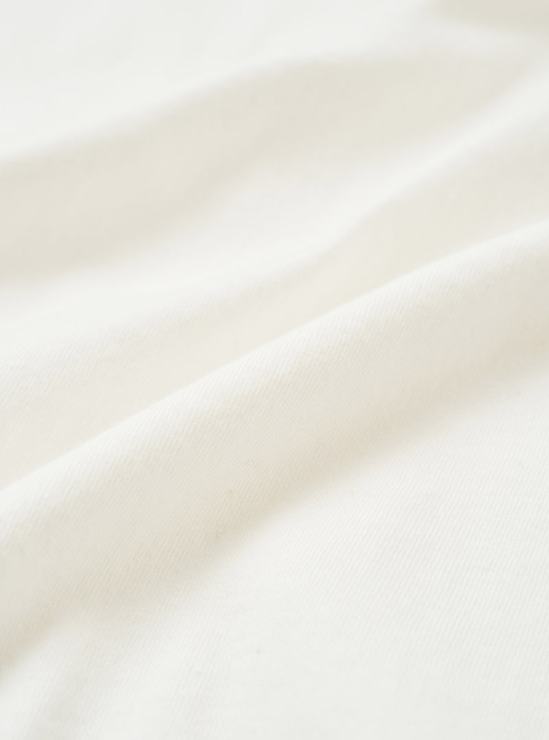 Close-up of ecru single jersey fabric, showing a soft, fine knit texture with natural drape and gentle folds. The off-white tone is even throughout, with a smooth surface and subtle sheen visible across the raised folds of the cloth.