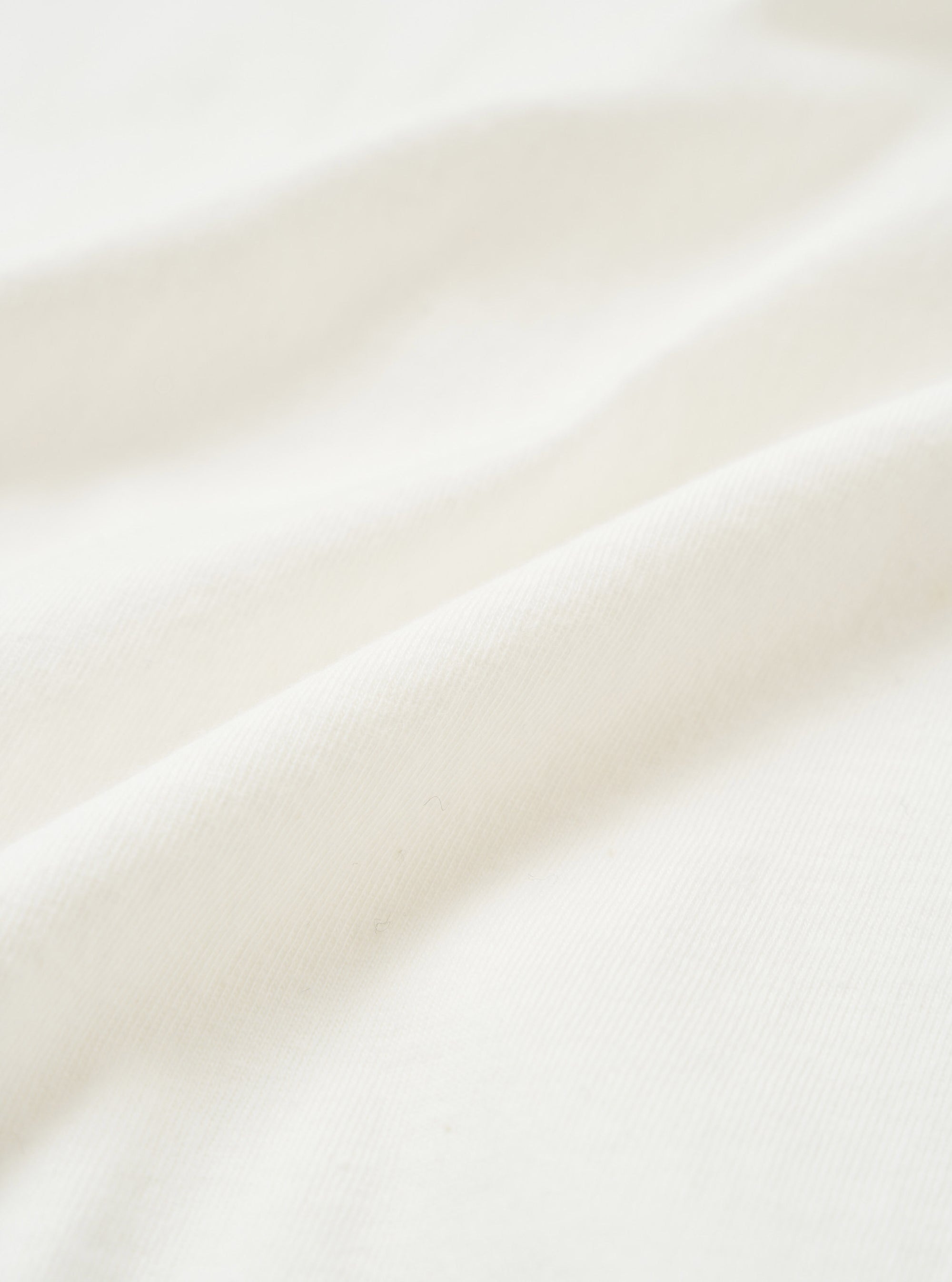 Close-up of ecru single jersey fabric, showing a soft, fine knit texture with natural drape and gentle folds. The off-white tone is even throughout, with a smooth surface and subtle sheen visible across the raised folds of the cloth.