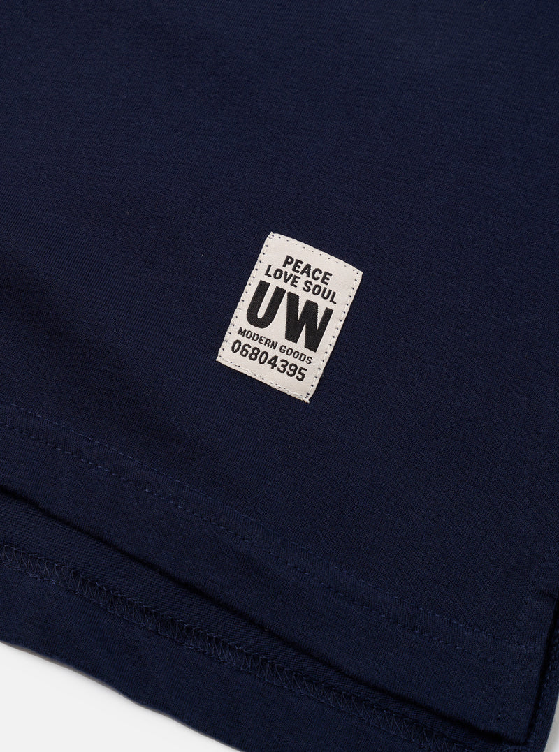 Close-up detail of a navy single jersey long-sleeve tee, flat lay on a white surface. The hem and topstitching are visible at the bottom of the frame. A woven rectangular label reads "Peace Love Soul UW Modern Goods 06804395" in black text on an off-white ground, stitched onto the chest.
