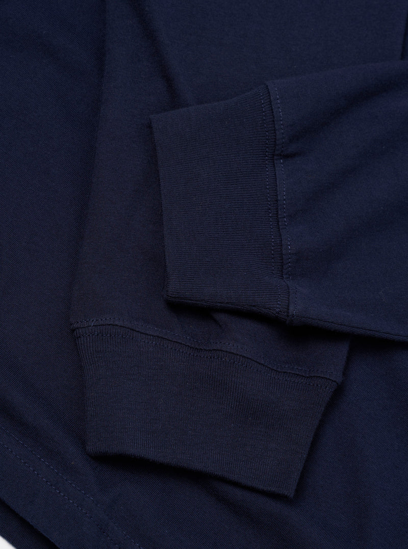 Close-up flat lay of a navy single jersey long-sleeve pocket tee. Two ribbed cuffs are prominent, showing overlock stitching along the seams. The smooth cotton jersey fabric is visible across the sleeve and body. A portion of a chest pocket with topstitching is visible at the bottom left.