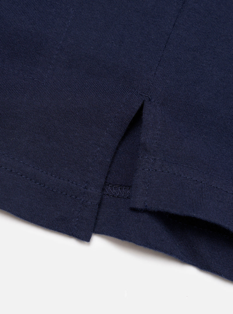 Close-up detail of a navy single jersey long-sleeve pocket tee, shot on a white studio surface. The image focuses on the curved hem and side seam vent, showing a twin-needle hem stitch and overlock seam construction on smooth, fine-knit cotton jersey fabric.