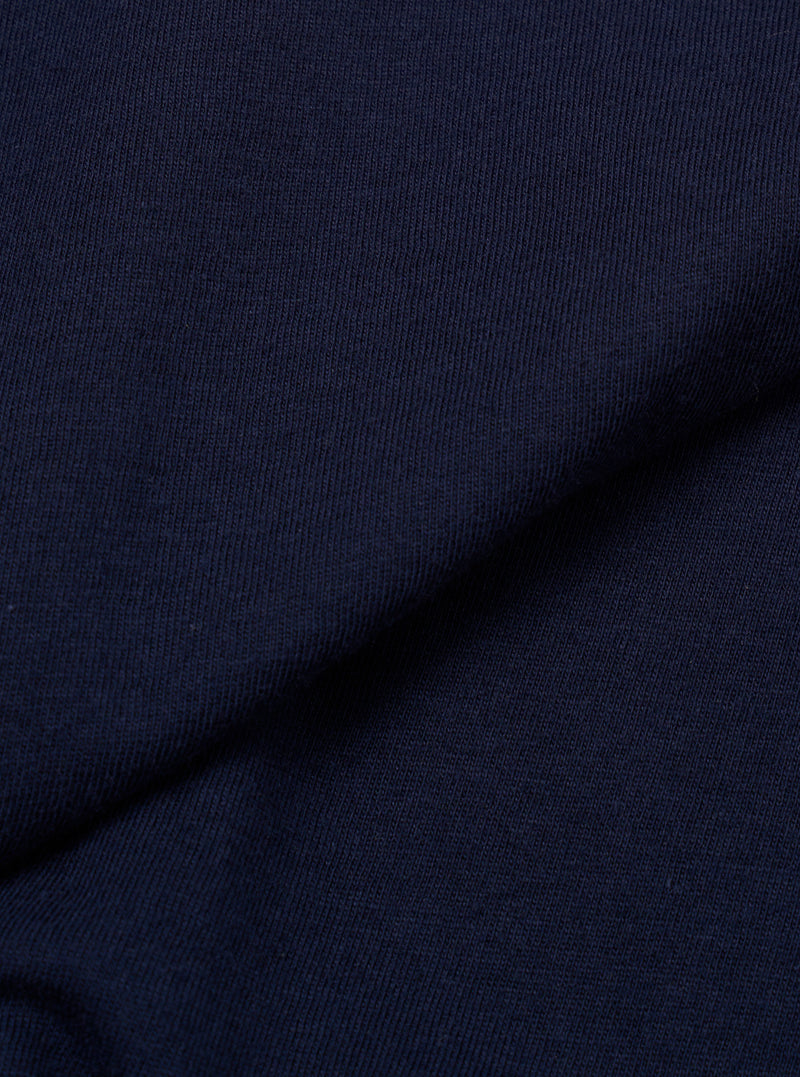 Close-up of navy single jersey fabric showing a fine knit texture with subtle slub variation. A diagonal fold runs across the frame, creating depth and shadow. The plain, solid navy colour is consistent across the textile surface.