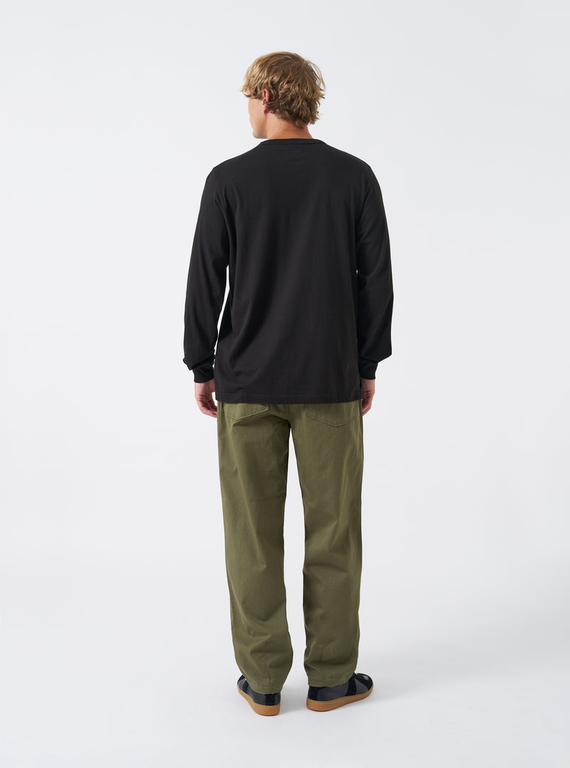 Back view of a model wearing the Black Single Jersey L/S Core Pocket Tee against a plain light studio backdrop. The image focuses on the reverse side of the black long-sleeve T-shirt, showing shoulder seams and clean hem.