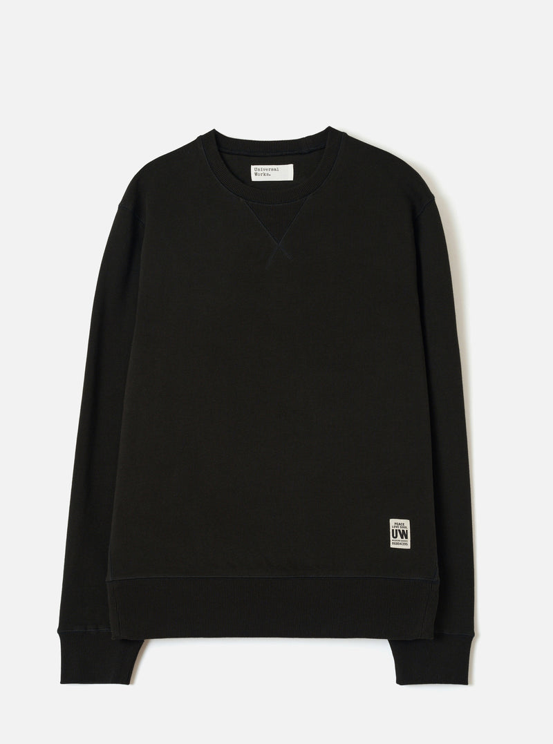 Black crew neck sweatshirt laid flat on a white studio backdrop. Ribbed collar with a V-stitch detail at the chest, ribbed cuffs and hem. Small woven UW brand label at the lower right. Universal Works woven label visible inside the collar. Smooth brushback jersey fabric throughout.