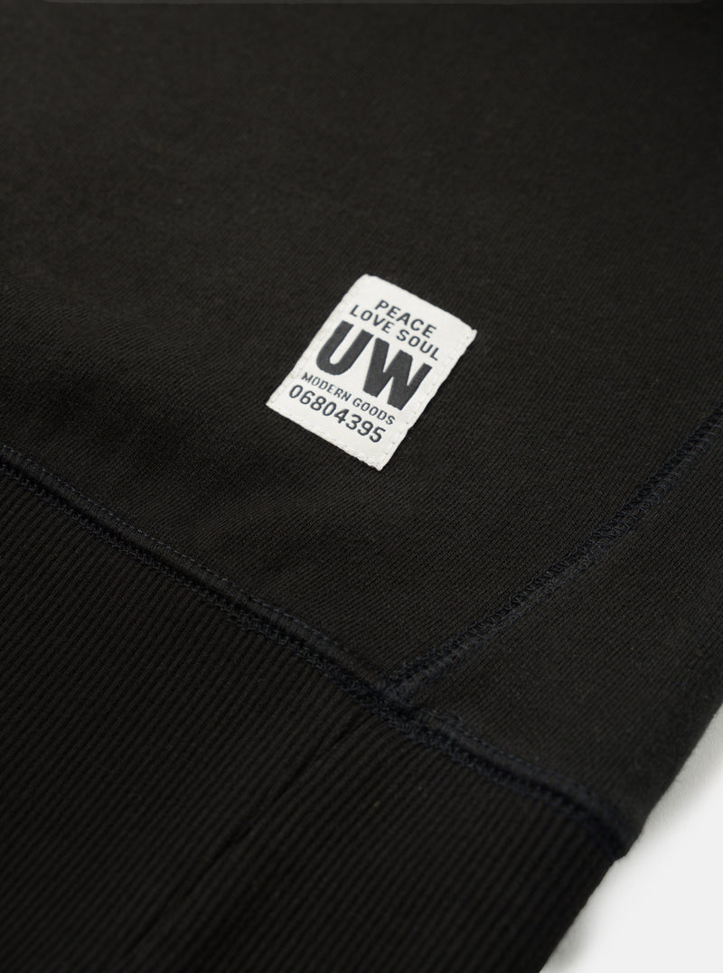 Close-up detail of a black brush back sweatshirt, shot flat on a white surface. The ribbed cuff is visible at the bottom, with topstitched seams where it meets the body. A woven square label reads "Peace Love Soul UW Modern Goods 06804395" in dark text on a white ground.