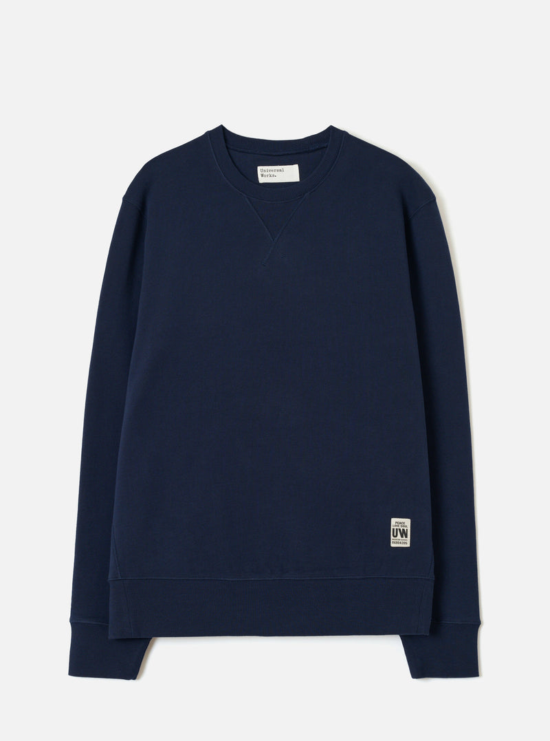 Navy brushback fleece crew-neck sweatshirt laid flat on a light grey studio backdrop. Ribbed cuffs and hem band, small V-stitch detail at the neckline, and a Universal Works woven label at the neck. A small branded UW patch label sits at the lower right hem.