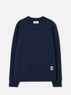 Navy brushback fleece crew-neck sweatshirt laid flat on a light grey studio backdrop. Ribbed cuffs and hem band, small V-stitch detail at the neckline, and a Universal Works woven label at the neck. A small branded UW patch label sits at the lower right hem.