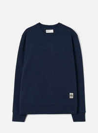 Navy brushback fleece crew-neck sweatshirt laid flat on a light grey studio backdrop. Ribbed cuffs and hem band, small V-stitch detail at the neckline, and a Universal Works woven label at the neck. A small branded UW patch label sits at the lower right hem.