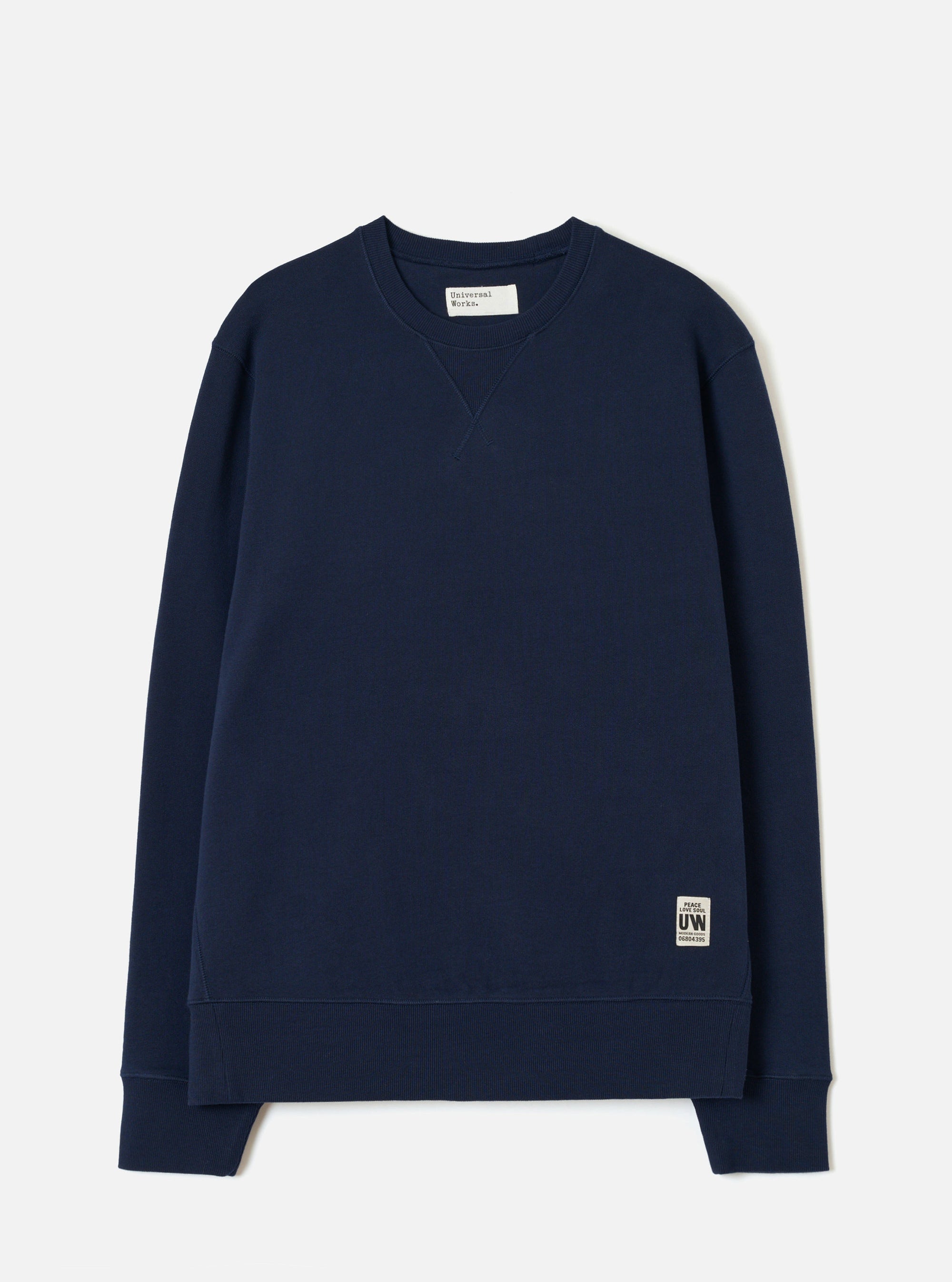 Navy brushback fleece crew-neck sweatshirt laid flat on a light grey studio backdrop. Ribbed cuffs and hem band, small V-stitch detail at the neckline, and a Universal Works woven label at the neck. A small branded UW patch label sits at the lower right hem.