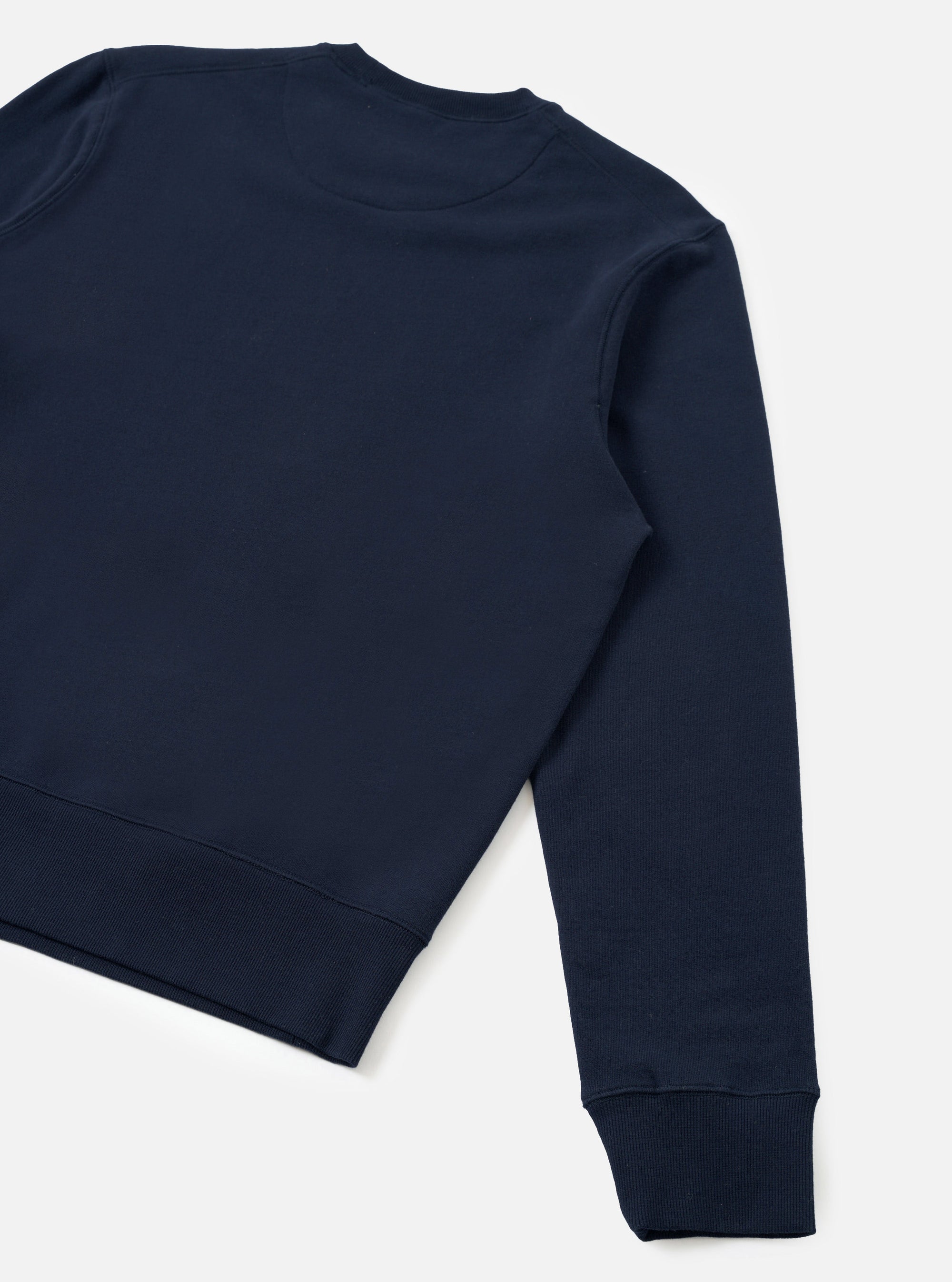 Navy brush back sweatshirt shot flat on a light grey seamless backdrop, angled to show the back and one sleeve. Crew neckline visible at top. Smooth jersey face on the body, one long sleeve extending to a ribbed cuff. Wide ribbed hem band at the base. Plain, no graphics or branding visible.
