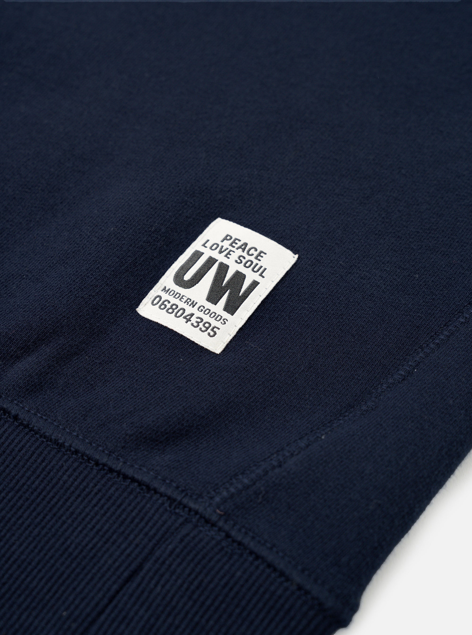 Close-up of the hem and cuff area of a navy brushback cotton sweatshirt, flat on a white surface. A woven off-white badge reads "Peace Love Soul UW Modern Goods 06804395". The ribbed cuff is visible at the lower left, with neat hem stitching running across the bottom edge.