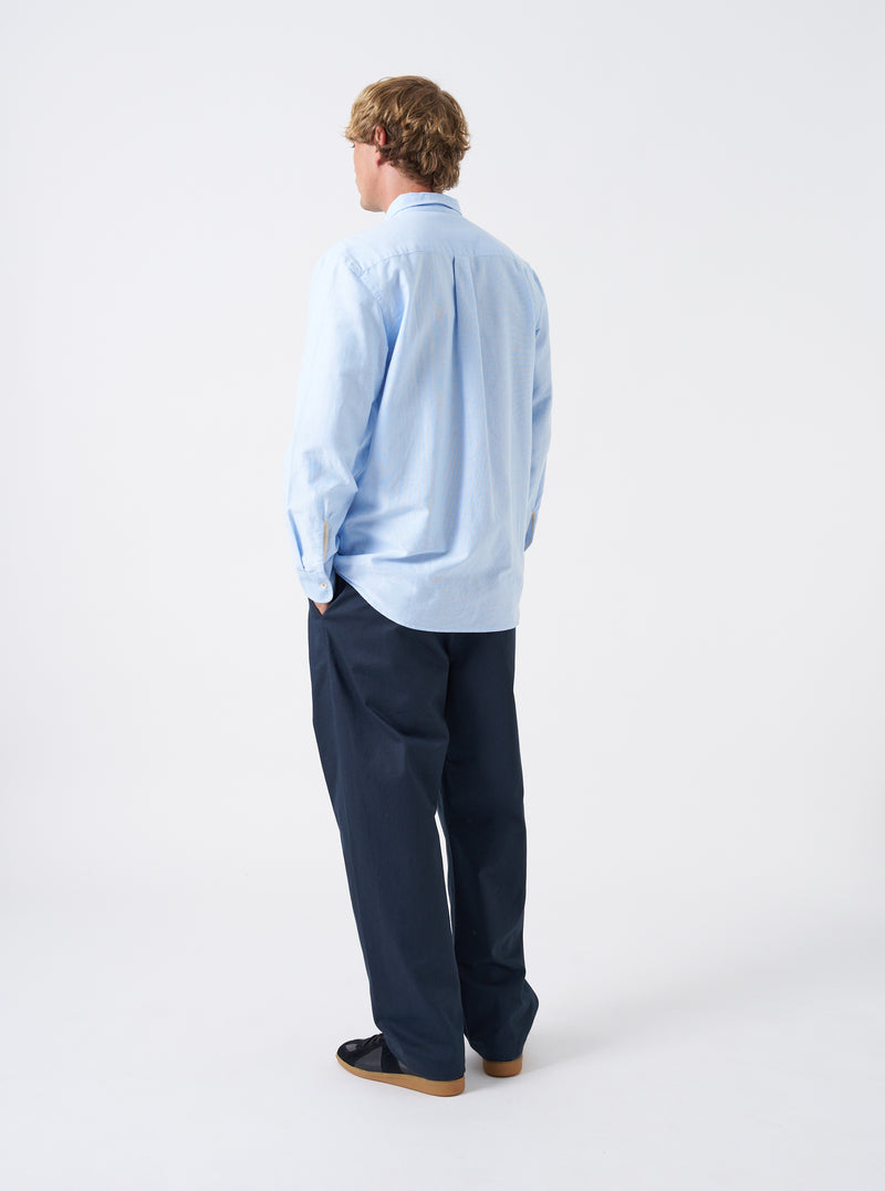 Angled view of a model wearing the Sky Blue Oxford Daybrook Shirt against a plain studio background. Highlights the side profile fit, sleeve drape, cuff finish, and classic casual look of the organic oxford garment.