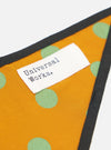Close-up of the Mustard Bold Dot Print Rayon Scout Scarf on a white background. The scarf is mustard with green dots and a black border. A white tag with black text is sewn on.