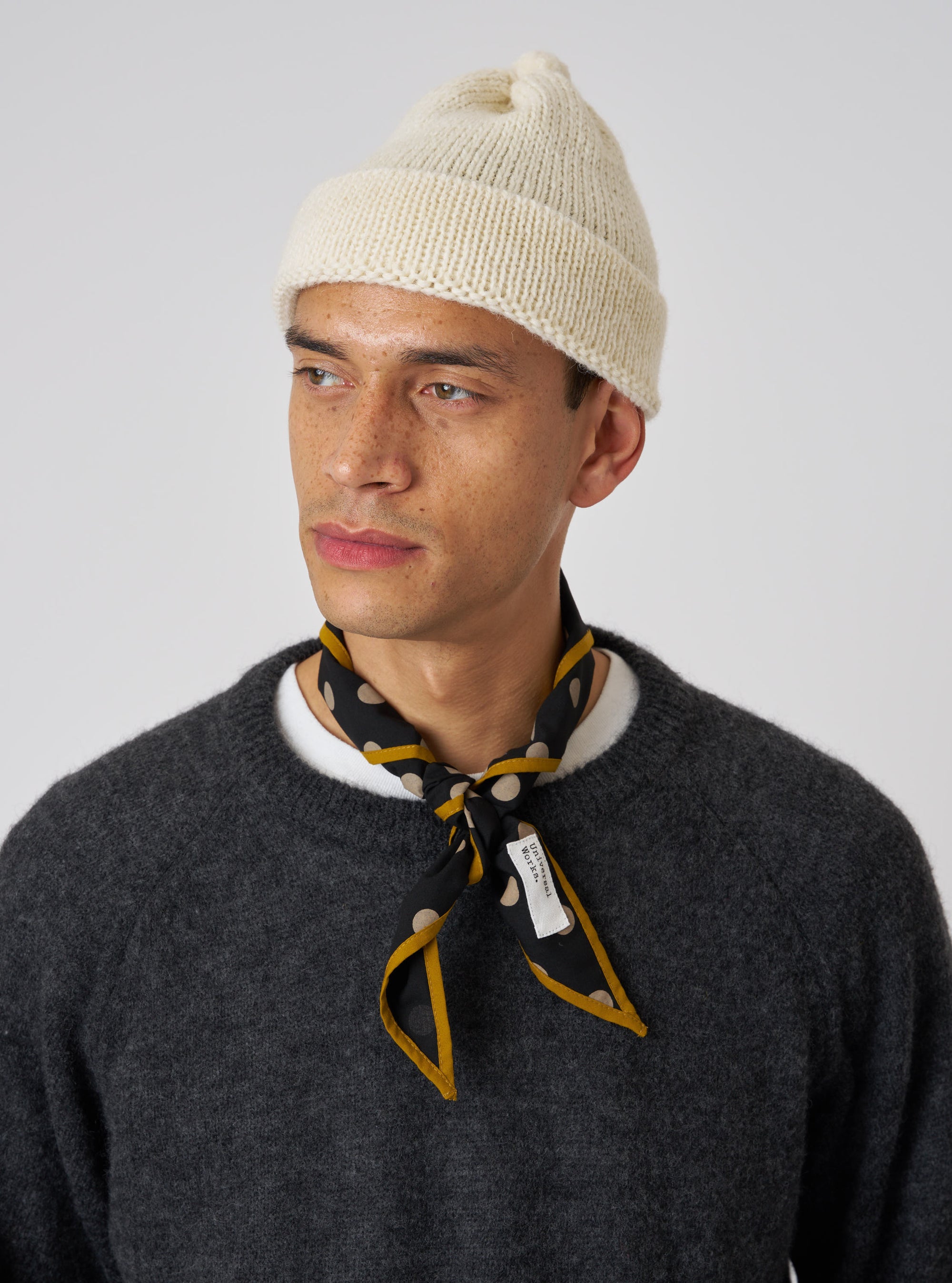 A model wears a Black Bold Dot Print Rayon Scout Scarf tied around his neck. The scarf features gold trim and is styled with a cream beanie and gray sweater against a white background.