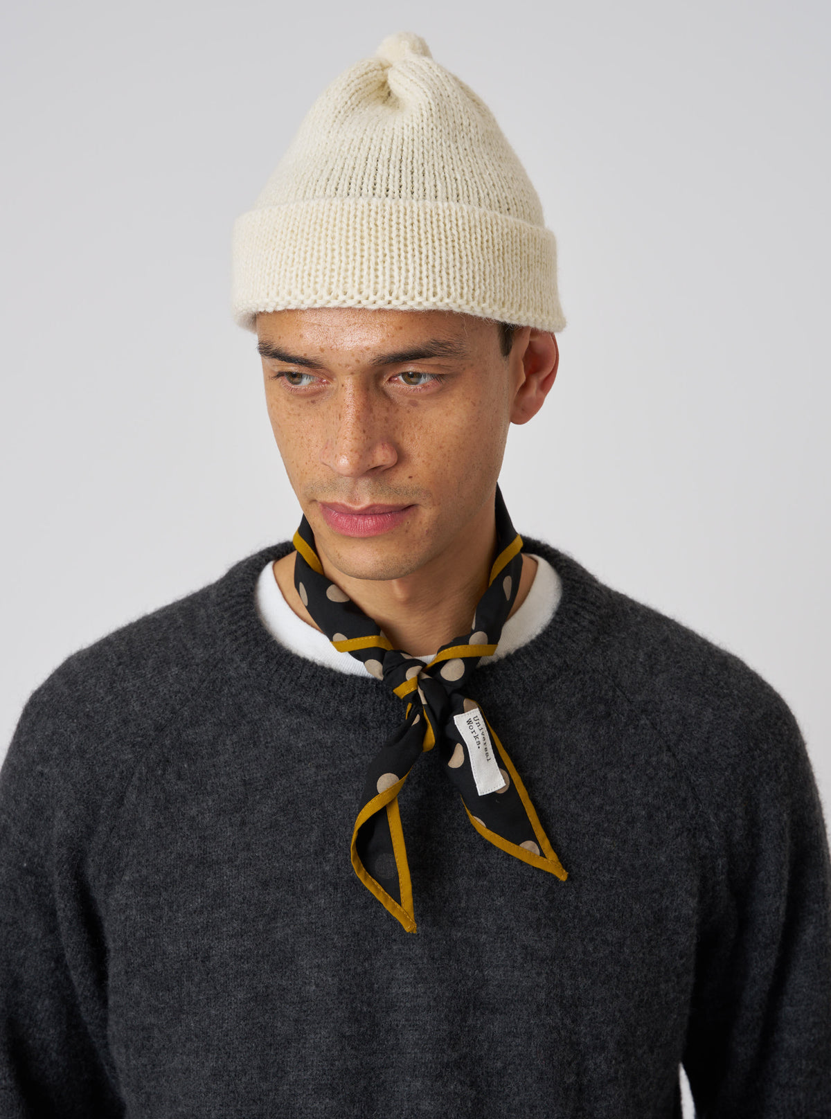 A model wears an Ecru Alpaca/British Wool Bobble Hat against a white background. The knitted hat is paired with a grey sweater and patterned neck scarf.