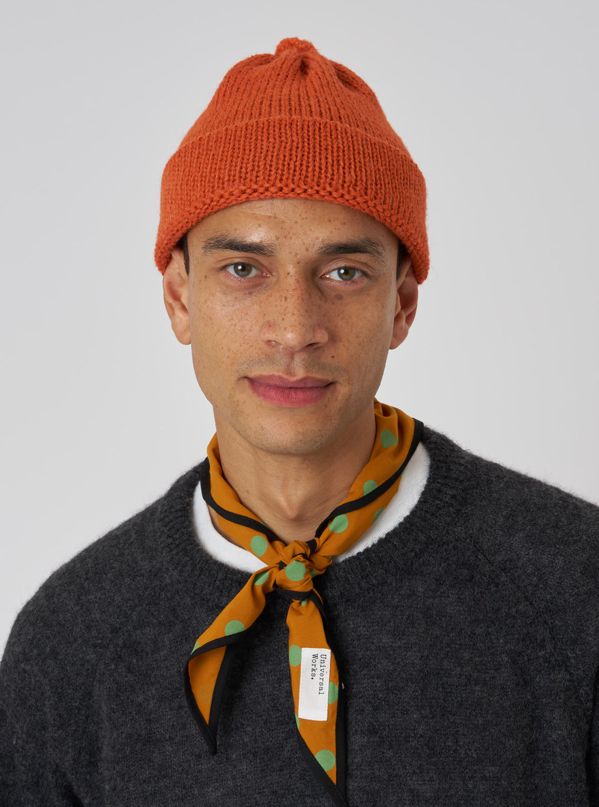 A model wears a Mustard Bold Dot Print Rayon Scout Scarf tied around his neck. The scarf is mustard with green dots and black trim. He also wears a gray sweater and orange beanie against a white background.