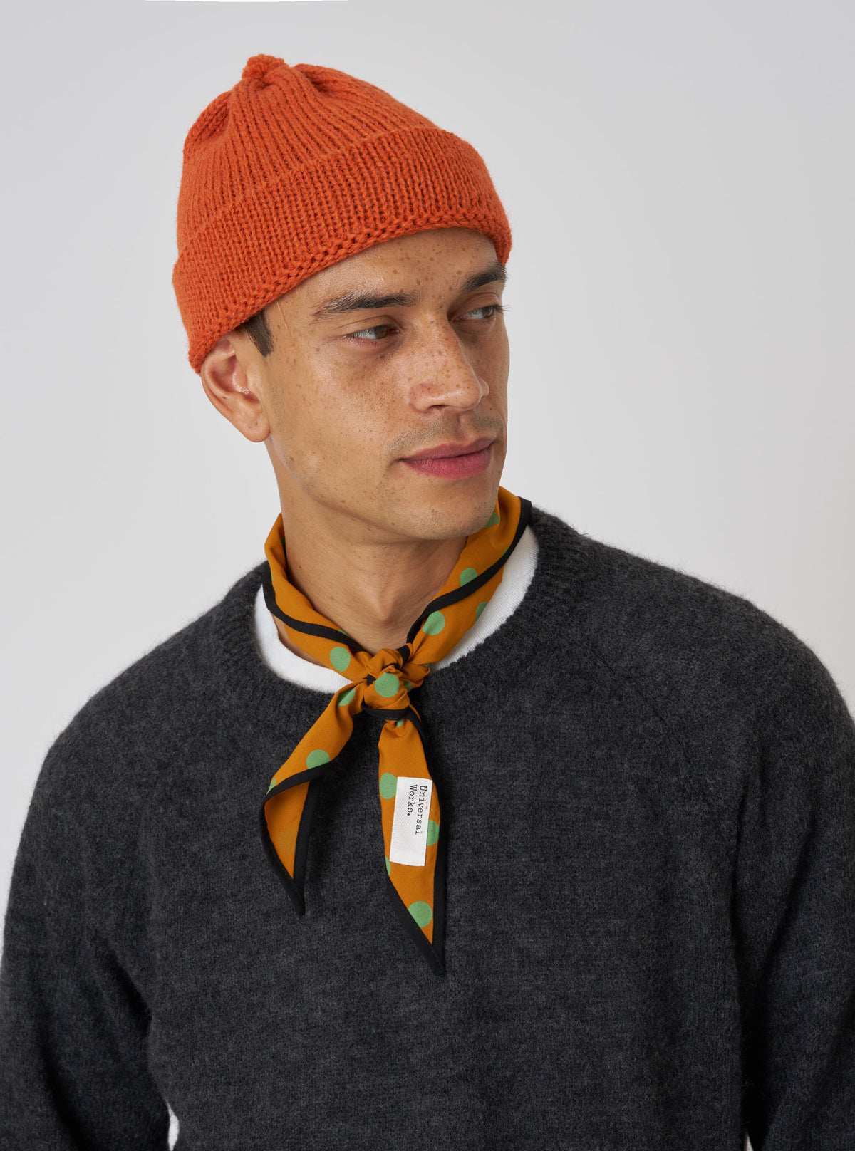 A model wears an Orange Alpaca/British Wool Bobble Hat against a white backdrop. The wool hat is paired with a grey sweater and patterned neck scarf.