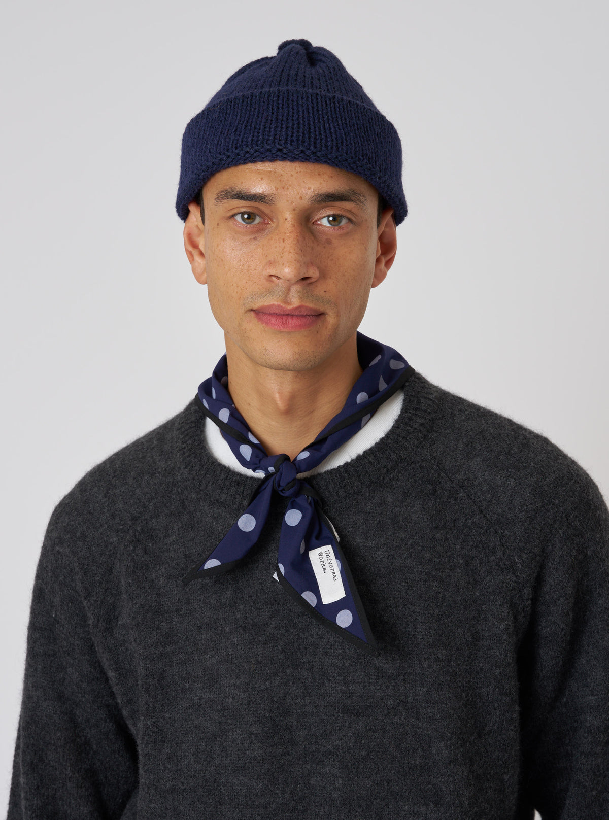 A model wears a Navy Alpaca/British Wool Bobble Hat against a white background. The hat is paired with a grey sweater and navy polka dot scarf.