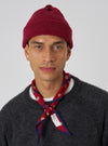 A model wears a Red Alpaca/British Wool Bobble Hat against a plain backdrop. The close-fitting hat is paired with a grey sweater and patterned neckerchief.