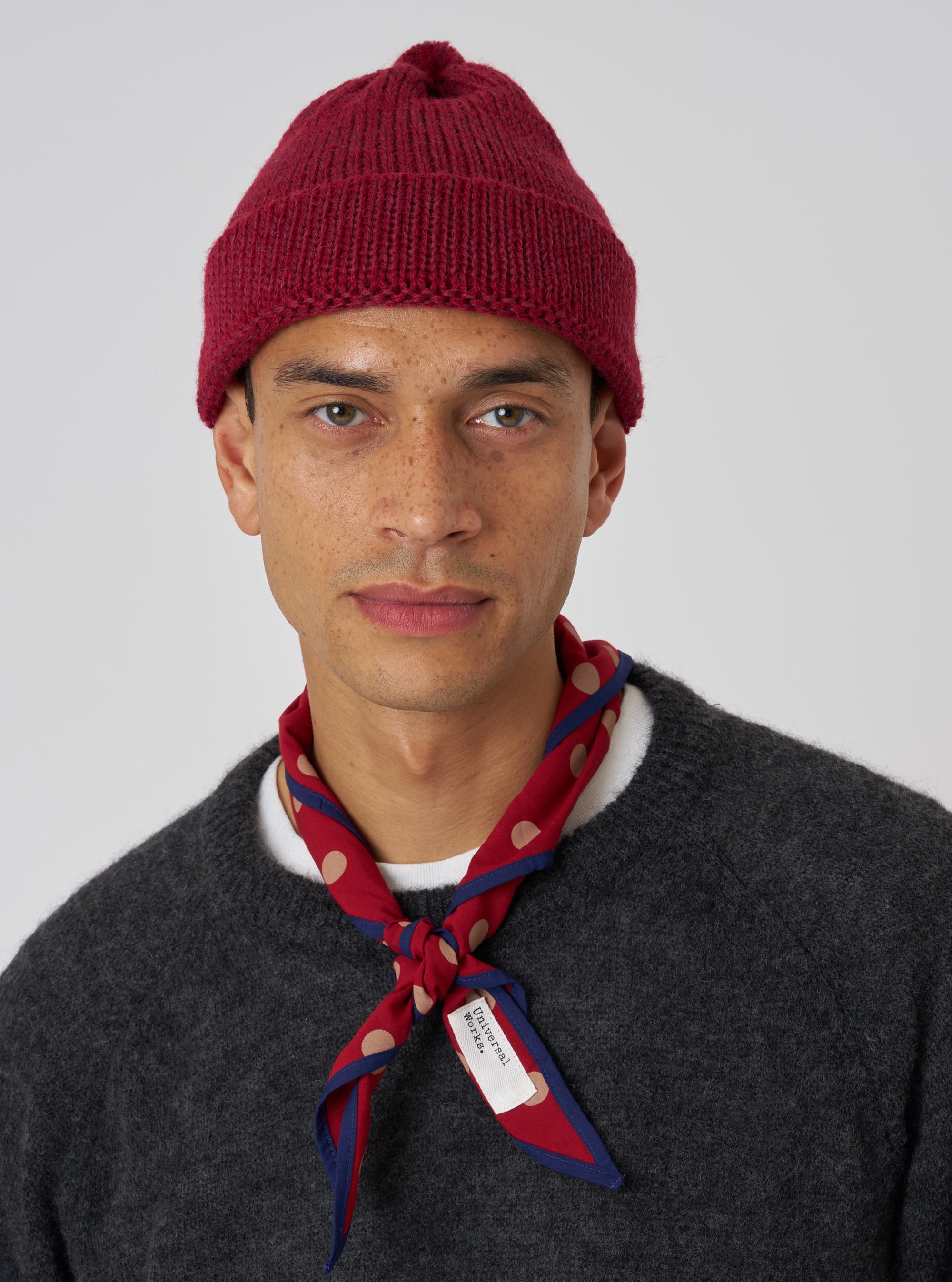 A model wears a Red Alpaca/British Wool Bobble Hat against a plain backdrop. The close-fitting hat is paired with a grey sweater and patterned neckerchief.