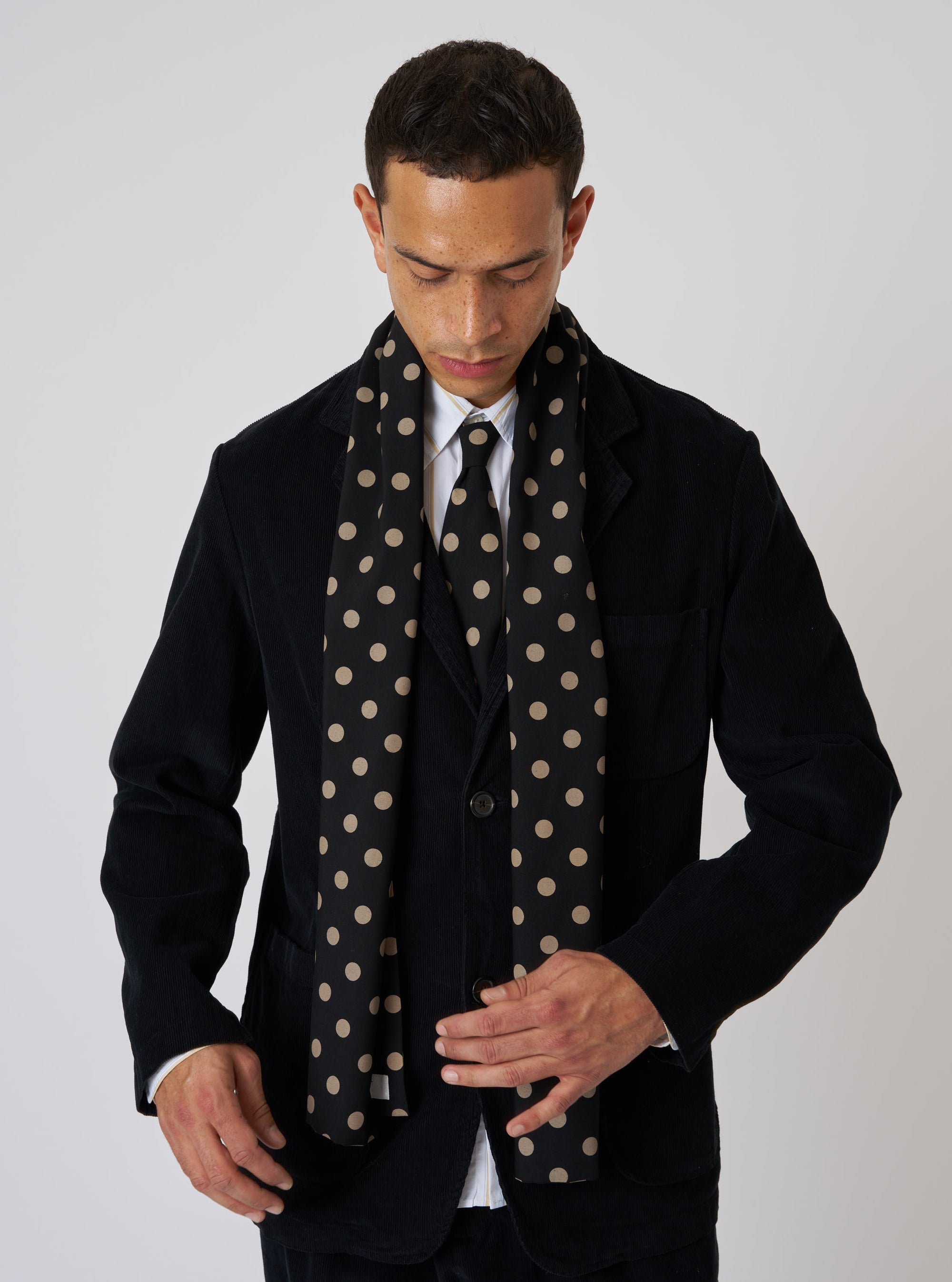 A model in a suit wears a Black Bold Dot Print Rayon Evening Scarf. The scarf has tan dots and is rayon. The background is a plain studio backdrop.