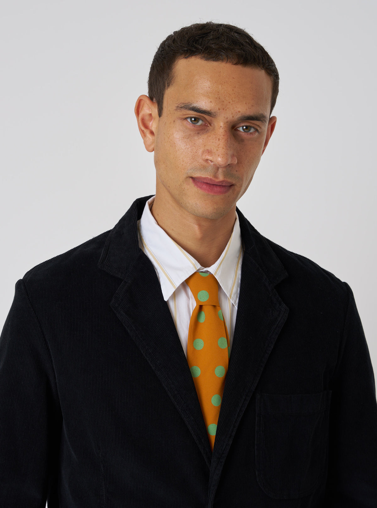 A model wears a Mustard Bold Dot Print Rayon Tie with a black corduroy jacket and striped shirt against a white backdrop. The tie is mustard with bold green dots.