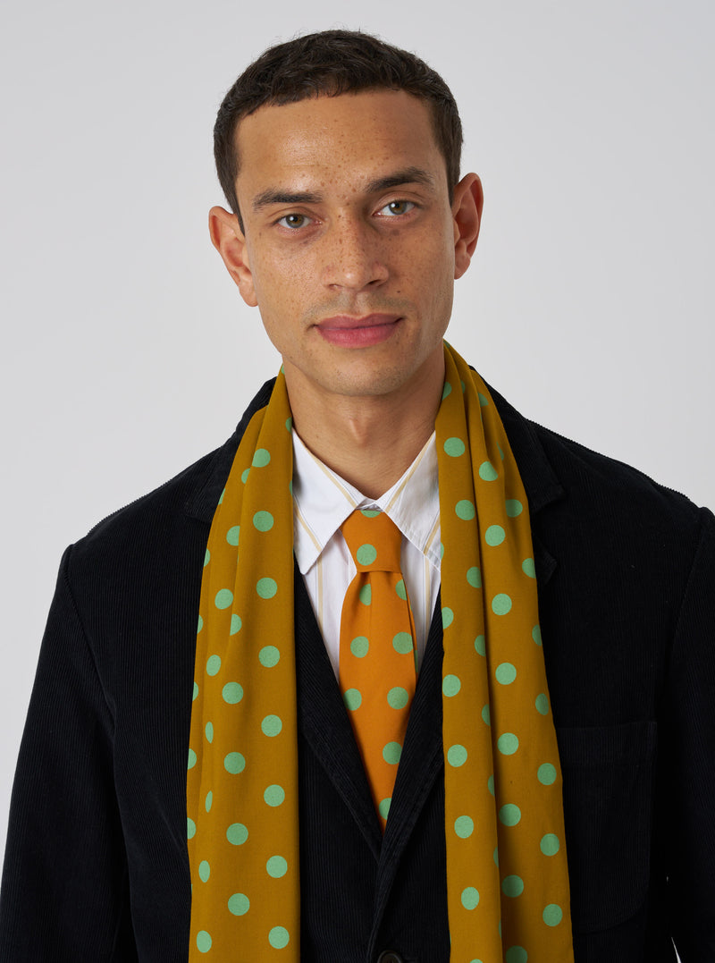 A model wears a Mustard Bold Dot Print Rayon Evening Scarf. The scarf is mustard with green dots. The background is a plain white wall.