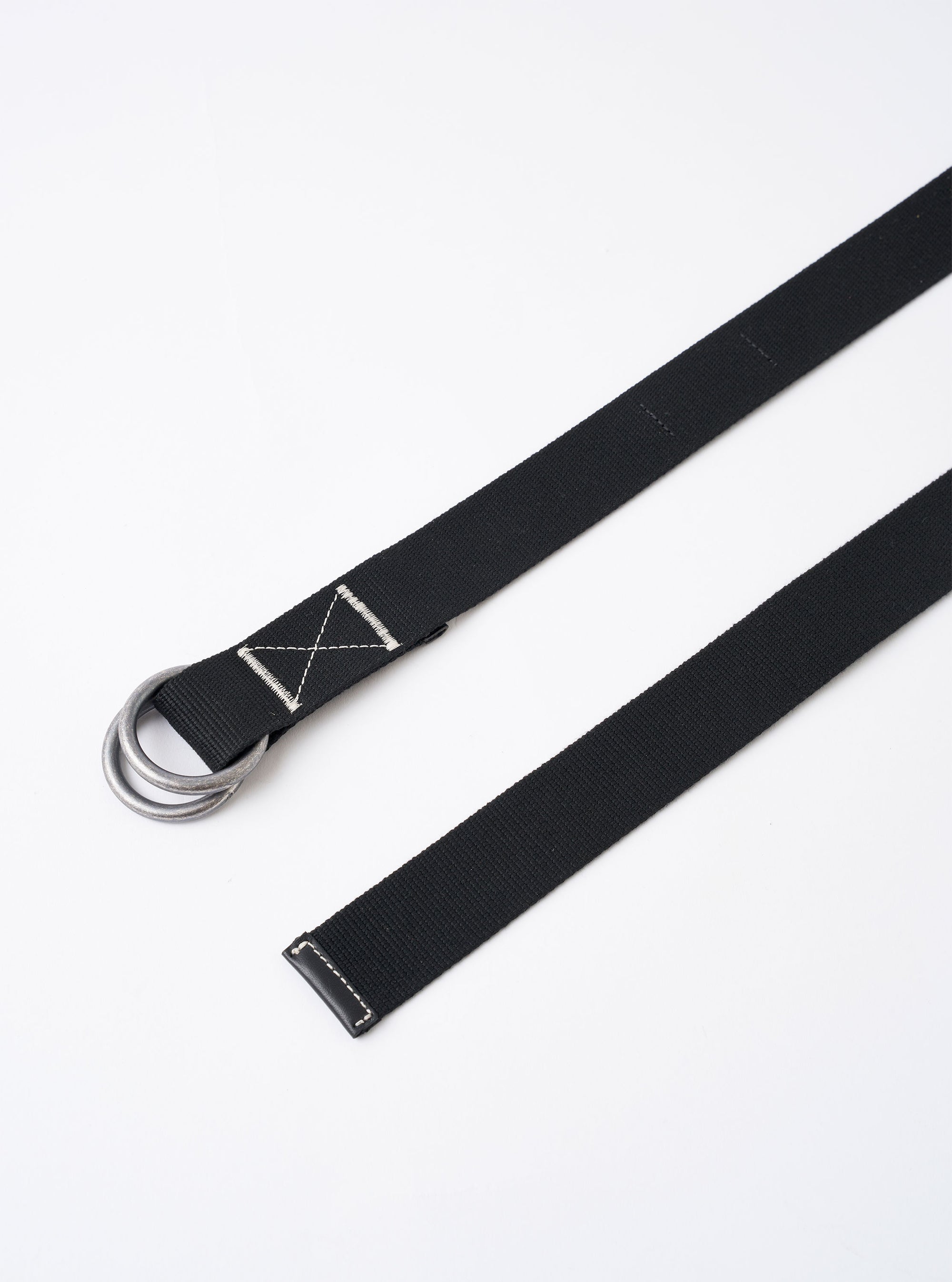 The Black Canvas Webbing O Ring Belt is shown as a flat lay against a white background. The belt features a black woven fabric strap and two silver metal O-ring buckles at one end. The belt is coiled to display its length and texture.