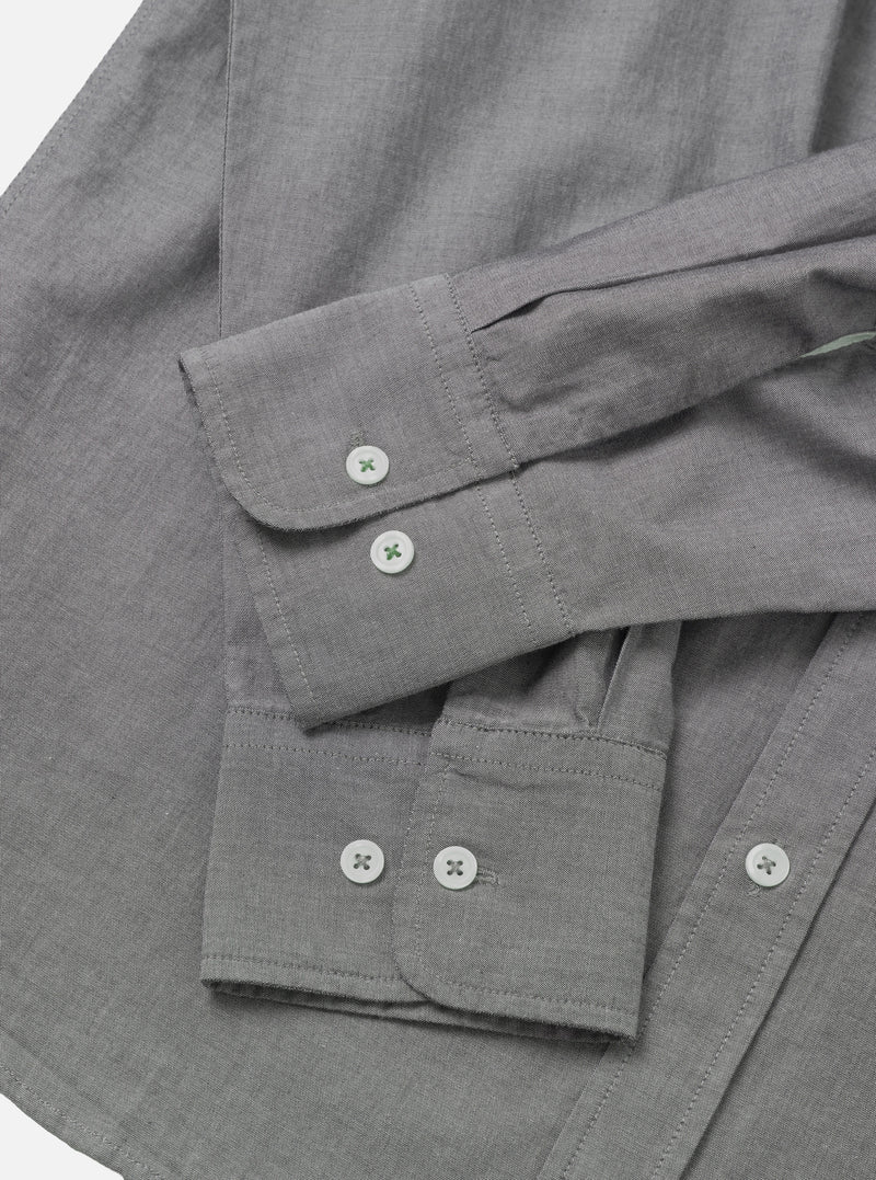 Close-up flat lay of a grey chambray shirt on a white surface, showing two barrel cuffs with white buttons stitched in green thread. The foreground cuff has two buttons side by side; the background cuff has one visible button. Top-stitching runs along the cuff edges. The fine chambray weave texture is visible across the body of the shirt.