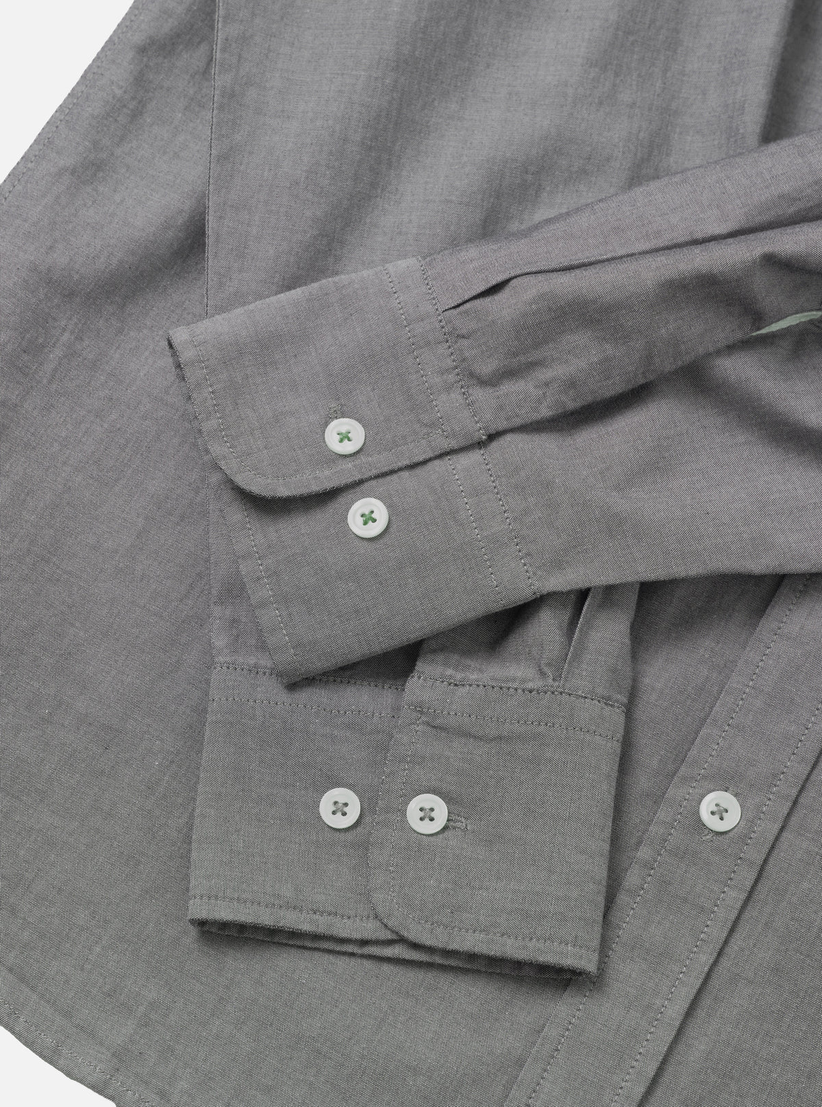 Close-up flat lay of a grey chambray shirt on a white surface, showing two barrel cuffs with white buttons stitched in green thread. The foreground cuff has two buttons side by side; the background cuff has one visible button. Top-stitching runs along the cuff edges. The fine chambray weave texture is visible across the body of the shirt.