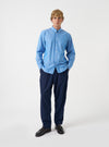 A male model poses against a plain studio backdrop wearing the Blue Prince Chambray Daybrook Shirt. The blue cotton chambray shirt features a button-up front, spread collar, and chest pocket.