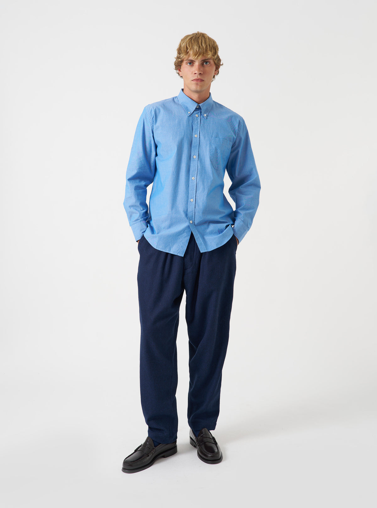 A male model poses against a plain studio backdrop wearing the Blue Prince Chambray Daybrook Shirt. The blue cotton chambray shirt features a button-up front, spread collar, and chest pocket.