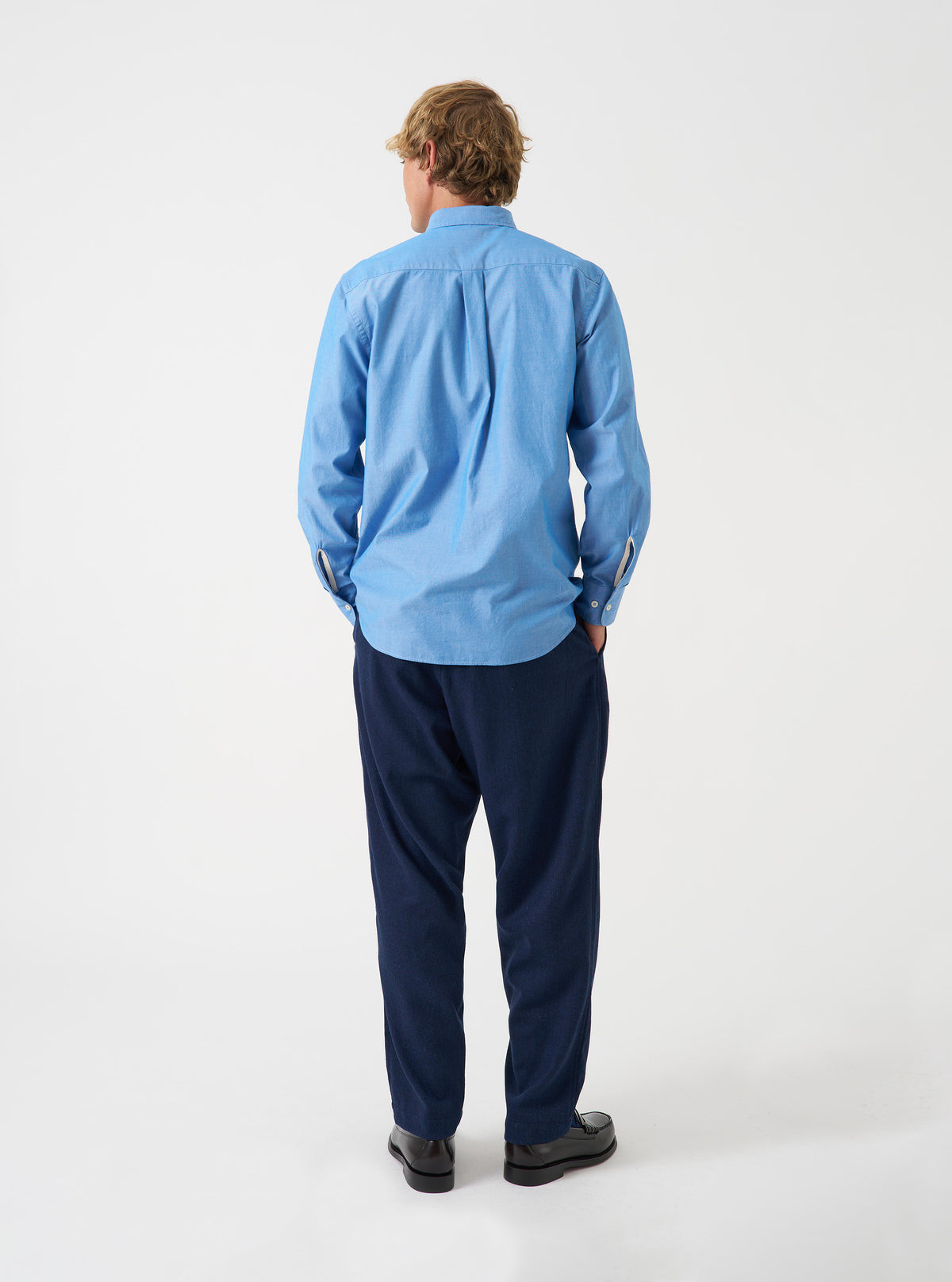 Back view of a male model wearing the Blue Prince Chambray Daybrook Shirt against a plain studio background. The blue chambray cotton shirt features a back yoke seam, long sleeves, and a straight hem.