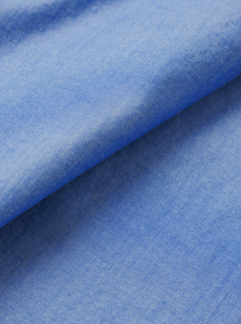 Close-up of the blue chambray fabric used in the Daybrook shirt, folded diagonally to show texture and drape. The plain-weave oxford-style cloth shows a fine two-tone weave of blue and white threads, giving a soft, mottled finish. Shot against a white studio backdrop.