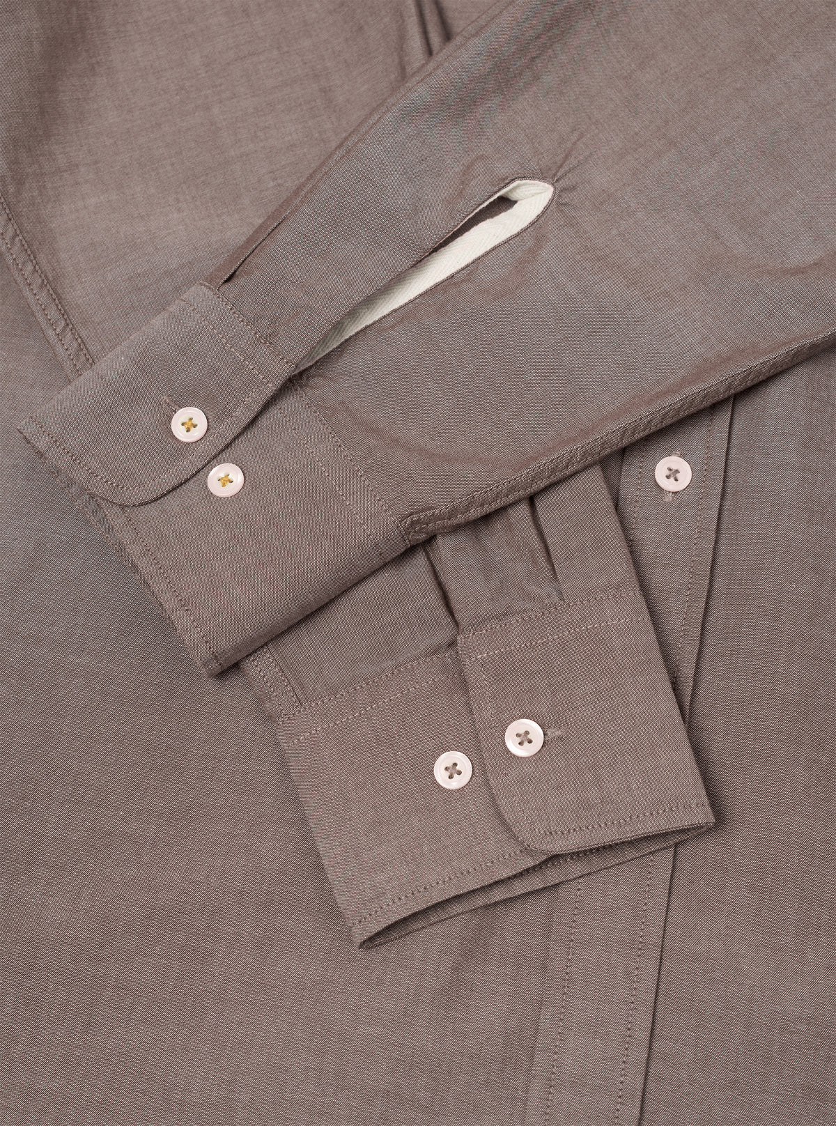 Close-up flat lay of a brown chambray shirt, focusing on the sleeve and cuff detail. Two barrel cuffs each fasten with two cream buttons. Topstitching outlines the cuffs and placket. A white contrast lining is visible inside the folded collar. The fine chambray weave is visible across the fabric.