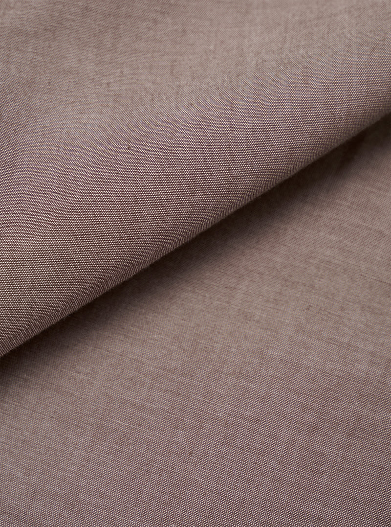 Close-up of the brown chambray fabric used in the Daybrook Shirt. The plain-weave textile shows a fine two-tone woven texture with subtle natural slubs across the surface. The fabric is folded diagonally, revealing depth and drape. No garment construction details are visible.