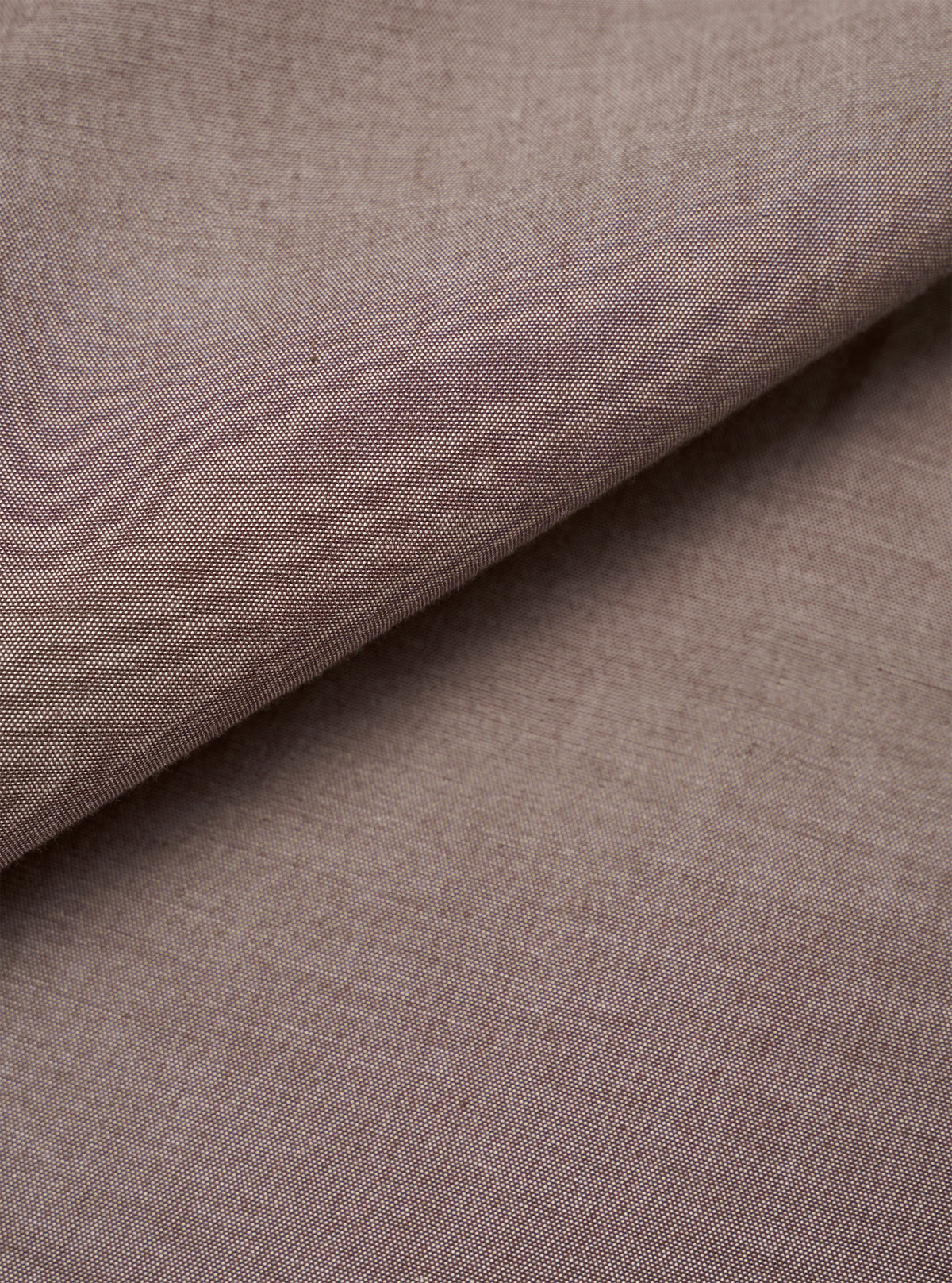 Close-up of the brown chambray fabric used in the Daybrook Shirt. The plain-weave textile shows a fine two-tone woven texture with subtle natural slubs across the surface. The fabric is folded diagonally, revealing depth and drape. No garment construction details are visible.