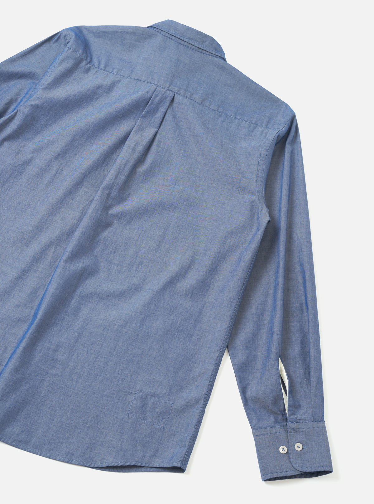 Rear flat lay of the Indigo Prince Chambray Daybrook Shirt on a light grey backdrop. The fine woven indigo chambray displays subtle two-tone texture, a back box pleat beneath the spread collar, and one long sleeve finished with a two-button cuff.