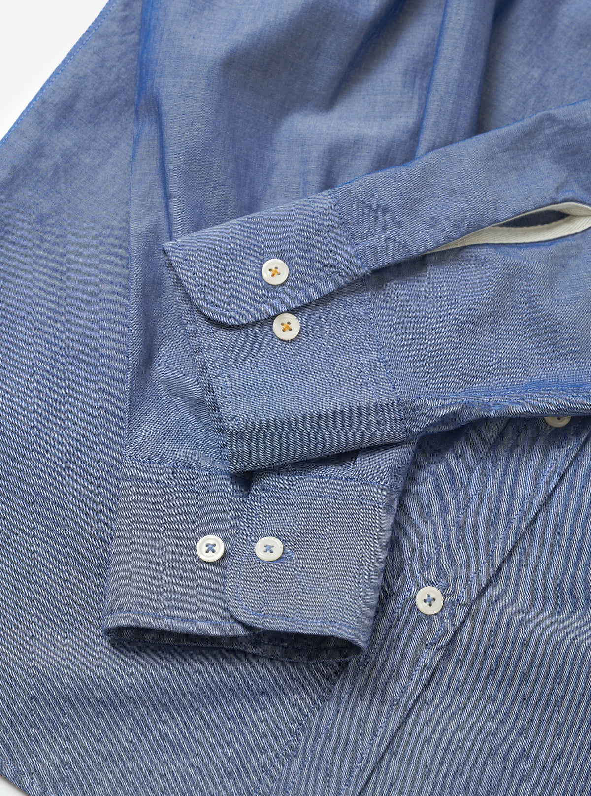 Close-up flat lay of the indigo chambray Daybrook shirt on a white surface, focusing on both cuffs. Each rounded cuff fastens with two mother-of-pearl buttons with coloured cross-stitching. The placket on the right shows two further buttons. Fine chambray weave texture is visible throughout, with a cream contrast lining visible inside one cuff.